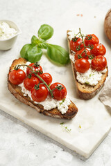 A sandwich with roasted cherry tomatoes with branch, fresh cottage cheese, green basil on a slice of whole wheat bread on a marble tray on grey background