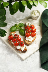 a sandwich with roasted cherry tomatoes with branch, fresh cottage cheese, green basil on a slice of whole wheat bread on a marble tray on grey background surrounded with green plant branches