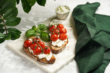 a sandwich with roasted cherry tomatoes with branch, fresh cottage cheese, green basil on a slice of whole wheat bread on a marble tray on grey background surrounded with green plant branches