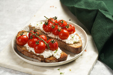 a sandwich with roasted cherry tomatoes with branch, fresh cottage cheese, green basil on a slice of whole wheat bread on a round plate on marble tray on grey background