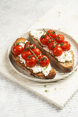 a sandwich with roasted cherry tomatoes with branch, fresh cottage cheese, green basil on a slice of whole wheat bread on a round plate on marble tray on grey background