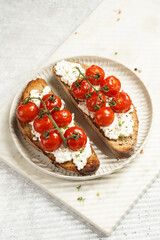 a sandwich with roasted cherry tomatoes with branch, fresh cottage cheese, green basil on a slice of whole wheat bread on a round plate on marble tray on grey background
