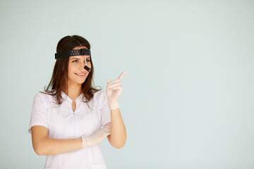 Cute young female otolaryngologist standing with illuminator on her head on bright background