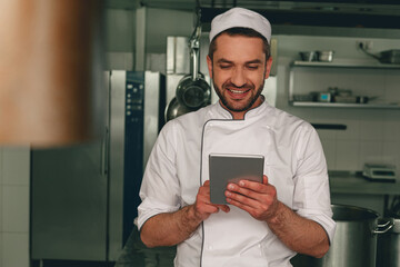 Smiling chef in uniform making order in digital tablet for the supplier standing on kitchen
