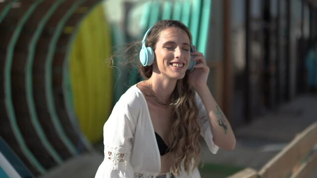 Chica joven de pelo largo escuchando m&uacute;sica con unos cascos celestes frente a una tienda de surf