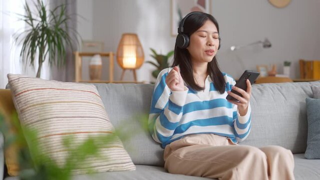 Serene asian lady in headphones listening favorite song with closed eyes while resting on cozy sofa in living room. Attractive woman holding smartphone and enjoying song.
