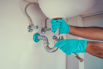 Plumber with blue gloves repairing sink siphon in kitchen. Plumber repairs and maintains chrome siphon under the washbasin. Refurbishment in the apartment. Elimination of leakage in the sink