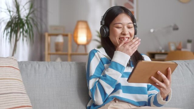 Happy cheerful asian woman wearing wireless headphones and watching comedy movie on modern tablet at home. Concept of relax time, technology and pleasure.