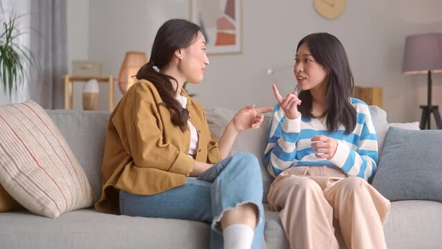 Young happy women in casual clothes gossiping about funny things and laughing at home. Asian female roommates spending free time together on cozy couch.