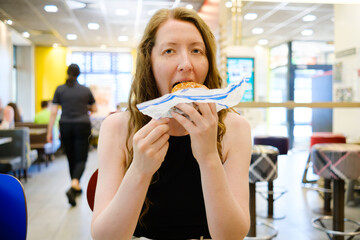Woman with burger in a fast food restaurant. Woman opening wrapping paper, holding, biting the burger and savouring food. Fast snack on the go