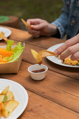 Salmon grilled fish with grilled vegetables in eco disposable tableware. Grilled potato, carrot, vegetable marrow. Man 
dipping potato in sauce. Vertical photo. Outdoor picnic in park on table. 