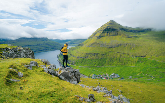 Male photographer stands on the edge of a cliff overlooking the Funningur fjord on Faroe Islands. Stunning view of the magnificent green cliffs and mountains in the picturesque Faroe Islands.