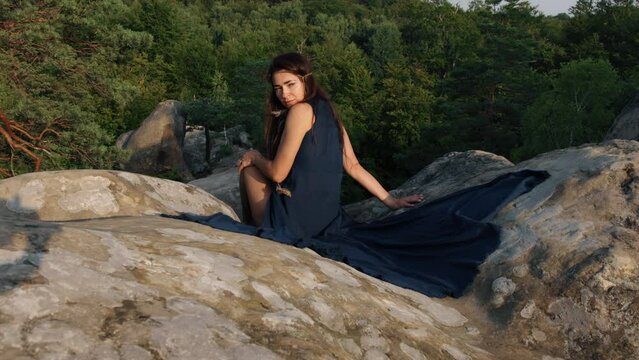 There is a young woman sitting on rocky terrain, relaxing outdoors while wearing a blue silk dress while enjoying a travel lifestyle in the background of mountains