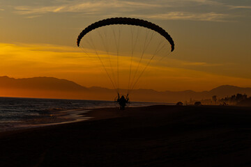 person in a motorised paraglider making a gliding flight on the shore of the beach at sunset or sunrise with an orange sky unrecognisable people
