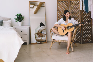 Brunette attractive female smiling and learning to play guitar while sitting in cozy home interior....