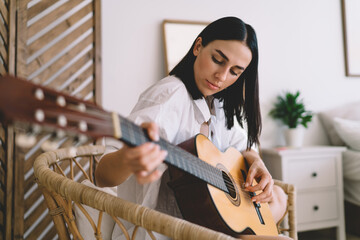 Brunette attractive female learning to play guitar while sitting in cozy home interior. Young...
