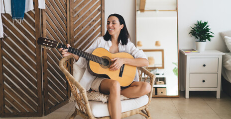 Brunette attractive female smiling and learning to play guitar while sitting in cozy home interior. Young happy woman enjoying her hobby with musical instrument performance relaxing on wicker chair