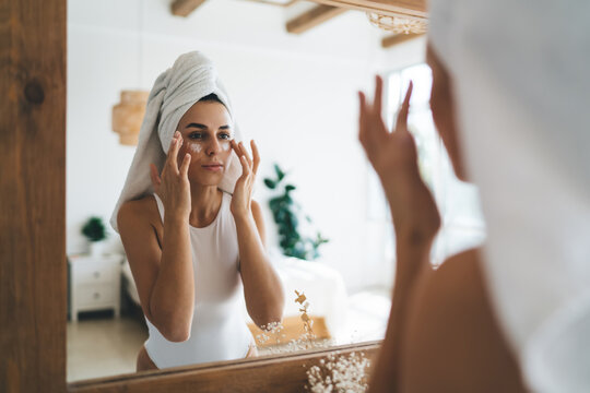 Young Beautiful Female With Towel On Head Looking To Mirror Reflection  Standing In The Bathroom At Home. 30 Years Old Woman Doing Daily Morning Rituals And Cleansing. Enjoying Healthy Skin Care