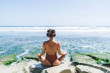 Back view of young female in swimsuit sitting in front ocean water meditating. Woman enjoying...