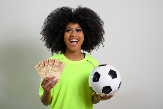 woman holding brazilian money is a soccer ball, she wears neon green uniform, isolated on gray background - Powered by Adobe