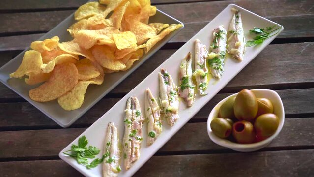 Three dishes with potato chips, picked anchovies and green olives on a dark wooden table with toothpicks. Typical Spanish snacks to eat in a bar. 