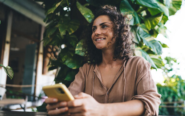 Cheerful female generation Z with modern mobile technology enjoying leisure pastime during weekend daytime, happy Caucasian woman holding smartphone device looking away and smiling outdoors