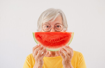 Cheerful elderly caucasian woman holding a slice of fresh watermelon in front of her mouth looking...