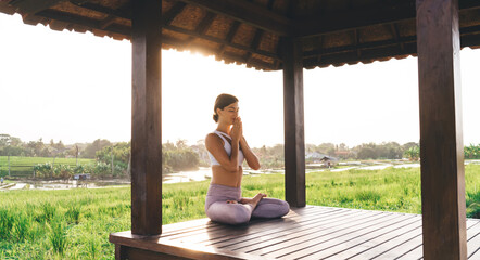 Side view of tranquil female in namaste praying near Indonesian rice fields and meditating for get...