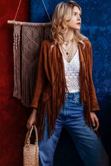 Fotobehang Boho Fashionable confident woman wearing stylish boho outfit with brown suede jacket with fringe, layered necklace, crochet top, jeans,  holding trendy round wicker straw bag, posing near colorful wall  © Victoria Fox