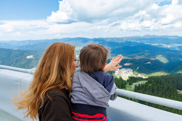 Mother holds son and shows him landscapes of forest valley of Rhodope Mountains while standing on tower of Snezhan