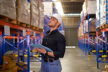 Young worker wearing helmet checking inventory and counting product on shelf in modern warehouse.