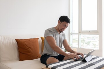 Angry and furious Asian trader man working on tablet or laptop in his apartment.