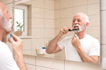 Senior man trimming and cutting beard using scissors and comb in front of the bathroom mirror