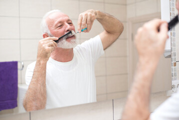 Senior man trimming and cutting beard using scissors and comb in front of the bathroom mirror