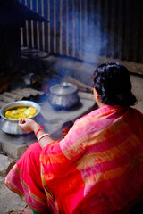 South asian village woman cooking in a mud made stove using wood and leaves as fuel, Bangladeshi hindu religious woman working hard wearing traditional dress 