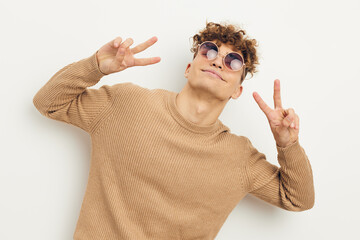 horizontal photo of a beautiful, happy man standing on a light background, wearing beige clothes and sunglasses, looking into the camera, showing a peace sign with his fingers