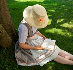 a woman in a beautiful traditional Bavarian dirndl dress (or Tracht) reading a book on the green...