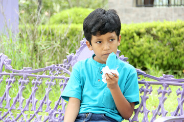 Poor Latino dark-haired boy with a blue t-shirt sitting on a park bench eating an ice pop because of the heat wave and cooling off in poverty by affecting his teeth with sweets and sugar
