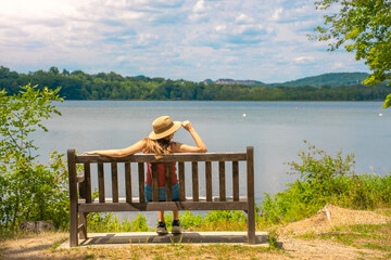 Back view of attractive young woman relaxing on a bench outdoors in front of a lake.