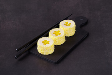Mousse cream dessert with lemon flower jelly, in the form of round sweet Japanese sushi, sprinkled with colored coconut flakes, on a serving board. Dark background