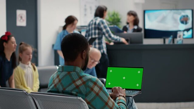 Young man holding greenscreen on laptop in hospital waiting room, sitting in lobby. Using copyspace display with isolated mockup template and blank chroma key background. Tripod shot.