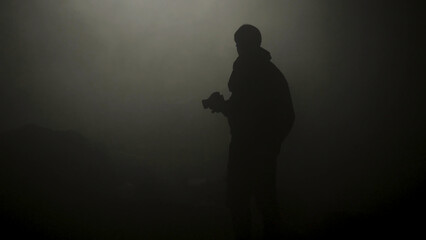 Black silhouette of man standing in smoky dark. Stock footage. Mysterious silhouette of young man stands in darkness lit only by dim light in haze