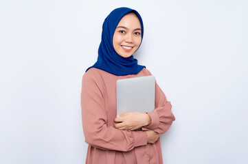 Smiling young Asian muslim woman in pink shirt holding laptop and looking at camera isolated over...