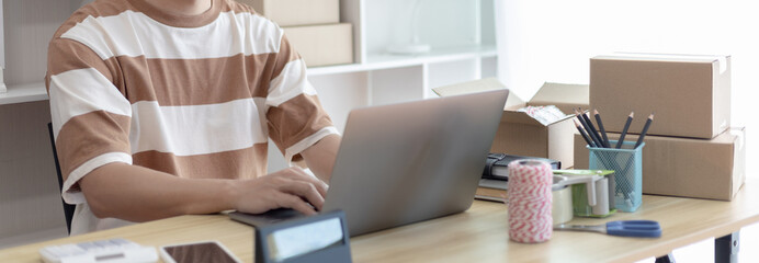 Young man uses a laptop to chat with customers who come to order product, Freelance work at home, Packaging on background, Sell online, Small business owner, Online shopping SME entrepreneur.