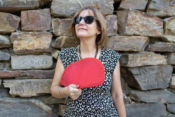 Portrait of a senior woman leaning on a stone wall outdoors and with a red fan due to the summer...