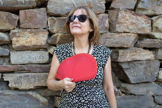 Portrait of a senior woman leaning on a stone wall outdoors and with a red fan due to the summer heat.