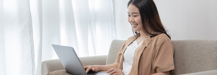 Asian woman sitting on the sofa with a laptop, Looking at laptop screen, Relaxing at home, Working through the Internet communication system, Happy lifestyle, Feel good.