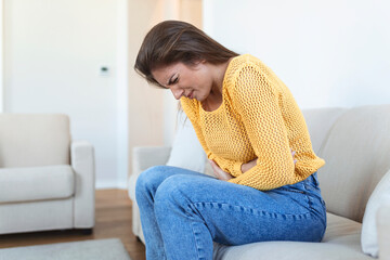 Obraz premium Woman lying on sofa looking sick in the living room. Beautiful young woman lying on bed and holding hands on her stomach. Woman having painful stomachache on bed, Menstrual period