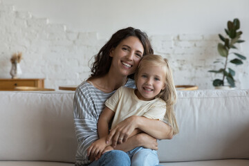 Happy joyful mom hugging happy sweet little preschool daughter, holding kid in arms, sitting on couch at home, looking at camera, smiling. Motherhood, family concept. Video call head shot portrait