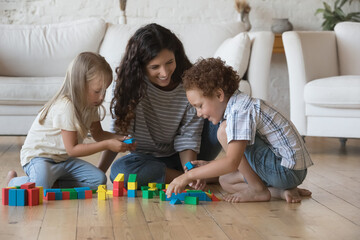 Happy friendly nanny, daycare teacher watching, entertaining children at home. Caring mom and cute little son and daughter kids playing wooden toy blocks on heating floor, enjoying learning game